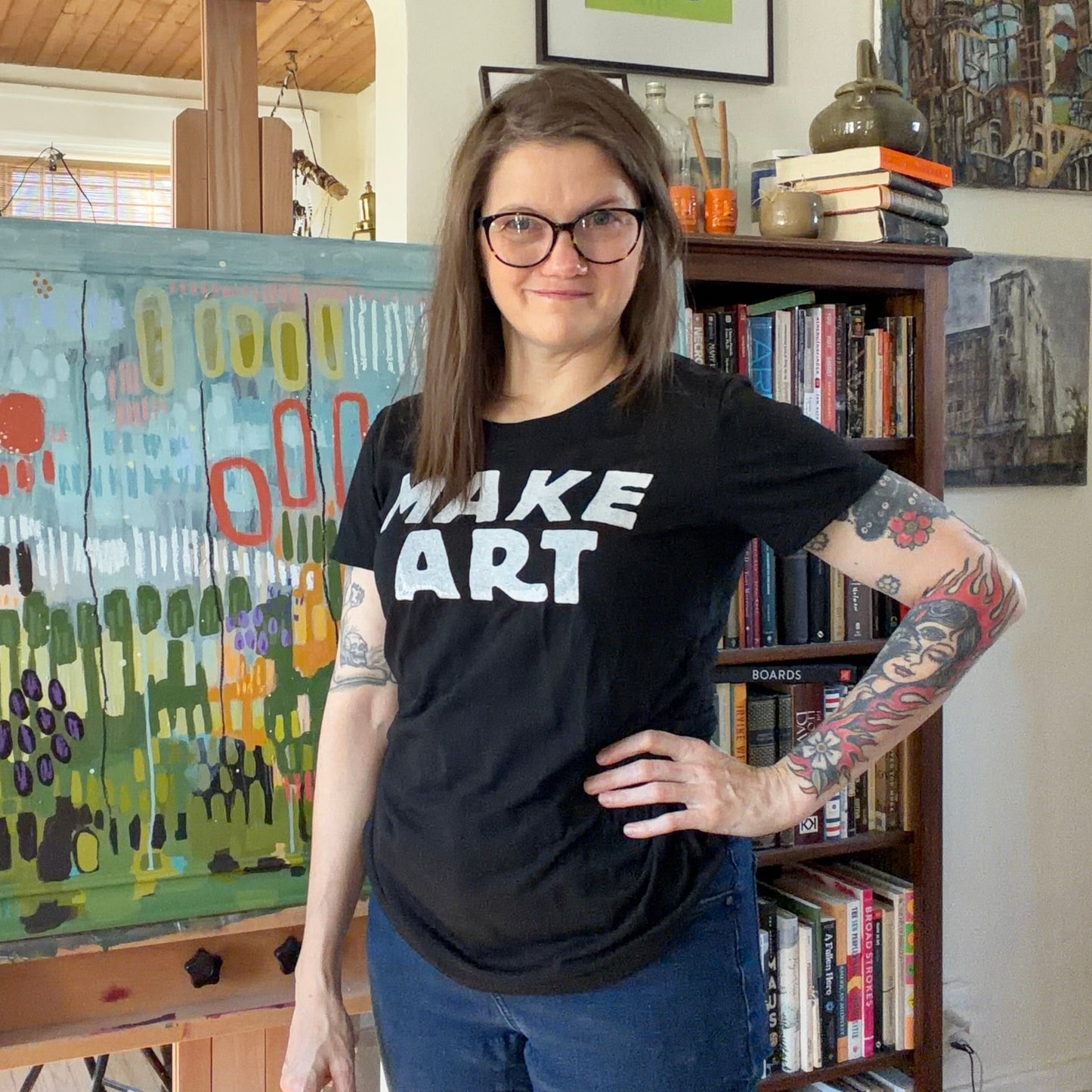 Person wearing a 'MAKE ART' t-shirt standing next to a colorful painting in an art studio.